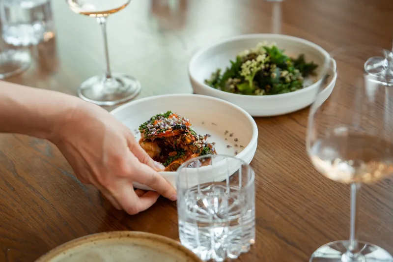 A server setting a shared course on the table beside wine glasses at Sheep
